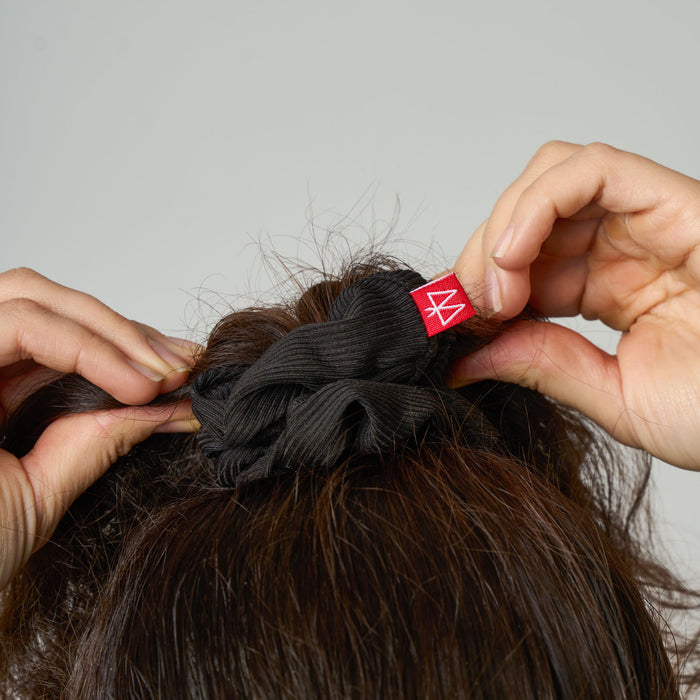 Person styling hair with a black scrunchie featuring a red logo on a neutral background