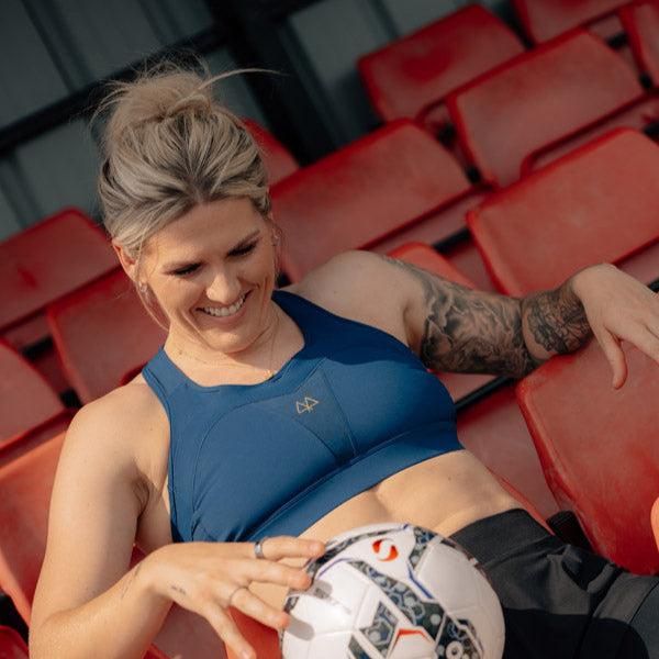 Chelsea Captain Millie Bright sitting on red stadium seats with a soccer ball, wearing a blue sports top and black shorts.