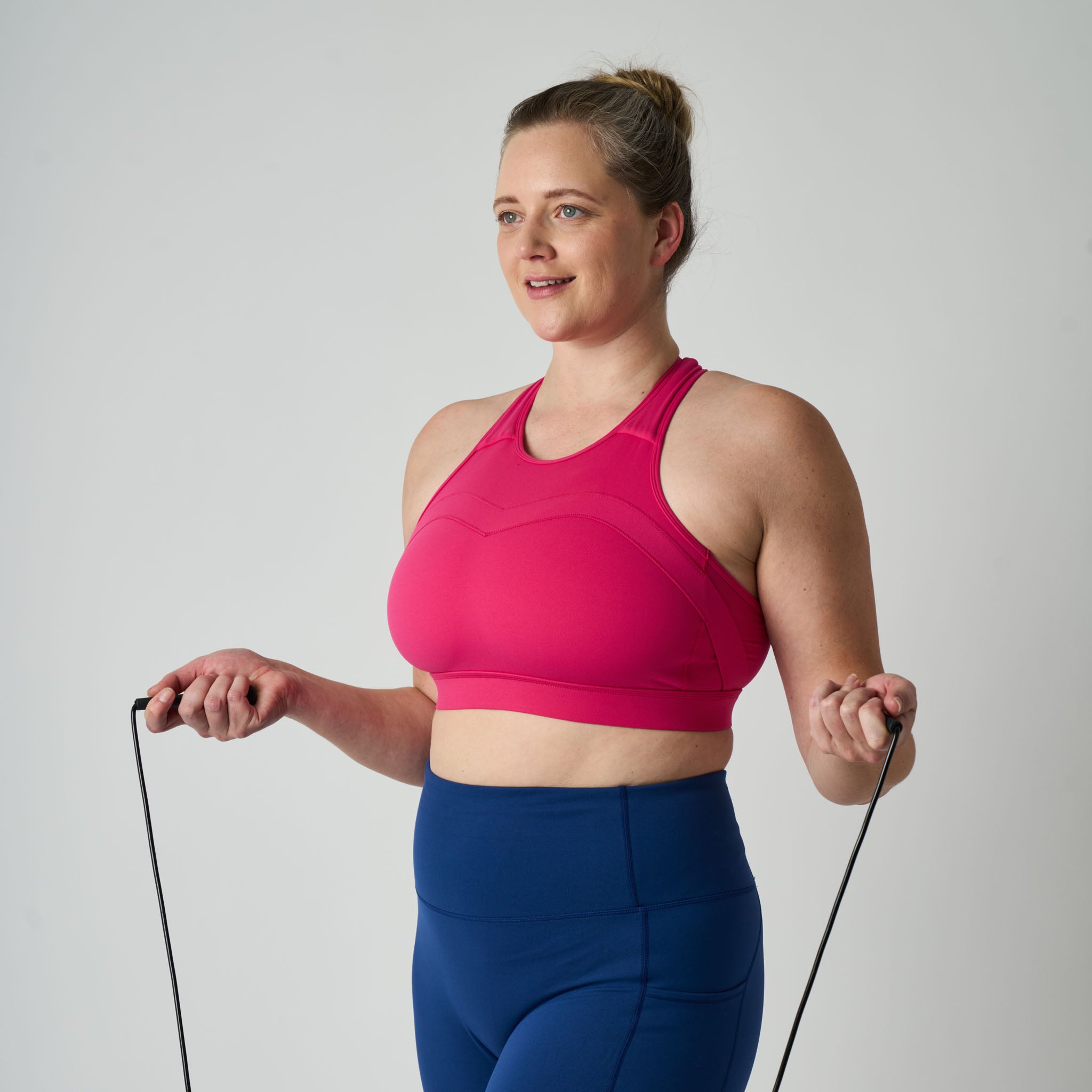 Woman in pink sports bra and blue leggings holding resistance bands against a plain background
